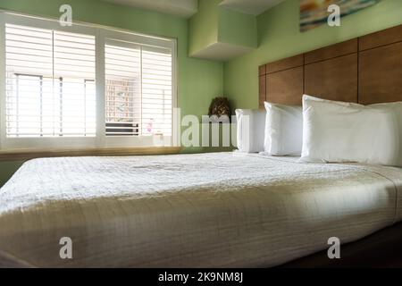 Camera da letto con luce naturale dalla finestra con cuscini bianchi, lenzuola e piumoni, trapunta sul materasso accanto alla testata del letto in legno nella moderna camera d'hotel Foto Stock