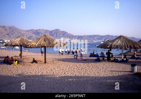 Scena della spiaggia ad Aqaba, Regno di Giordania, con alcune persone sdraiate all'ombra sotto gli ombrelloni di paglia e altri che godono il sole caldo Foto Stock