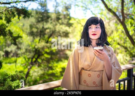 Ritratto di giovane ragazza caucasica donna felice volto sorridente, bevendo tazza di tè verde all'esterno nel giardino cortile esterno con capelli neri Foto Stock