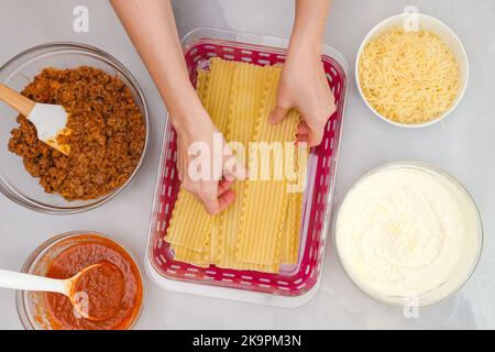 Tagliatelle di lasagna bollite, salsa di marinara, manzo macinato, formaggio in ciotole. Ingredienti per la ricetta di lasagne di manzo primo piano su tavolo da cucina, mani donna, piatto Foto Stock