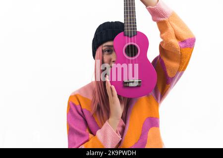 Concetto di passione musicale. Primo piano medio di una giovane ragazza caucasica dai capelli rosa in una beanie nera e cardigan arancione e rosa che tiene un nuovo ukulele rosa caldo in un modo che copre metà del suo volto. Foto studio con sfondo bianco, spazio copia. Foto di alta qualità Foto Stock