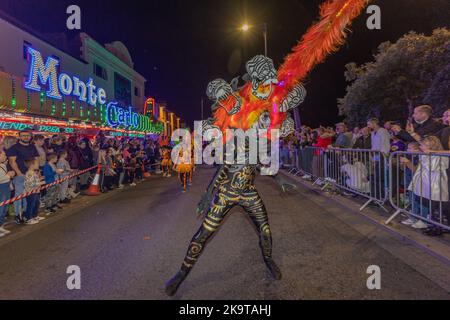 Southend on Sea, Regno Unito. 29 ottobre 2022. Southend Halloween Parade torna sul lungomare della città per un secondo anno. Un interprete mascherato in un drammatico costume nero e oro con piume rosse-arancioni danzano energicamente durante una vivace sfilata notturna. Gli spettatori fiancheggiano la strada dietro le barricate, illuminate da insegne al neon, tra cui "Monte Carlo". La scena cattura lo spirito festivo di una celebrazione pubblica. Penelope Barritt/Alamy Live News Foto Stock