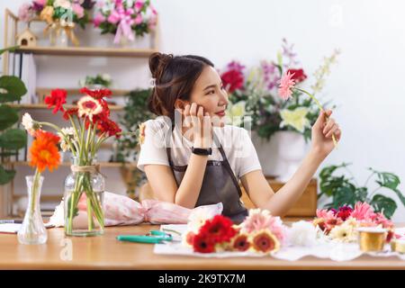 Concetto fiorista, fiorista femminile tocca mento sulla mano e sorriso mentre tiene su gerbera rosa in negozio. Foto Stock