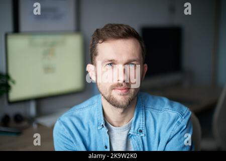 Allegro barbuto ingegnere software maschio seduto al posto di lavoro in ufficio guardando la fotocamera. Giovane uomo bello in camicia denim, emozione positiva con monitor per computer sullo sfondo Foto Stock