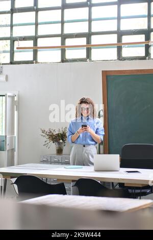 Felice giovane donna che usa il telefono cellulare in piedi in ufficio al lavoro. Foto Stock