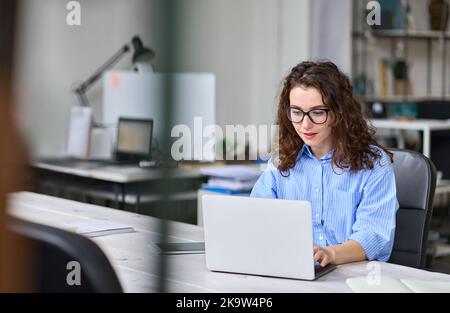 Una giovane donna in viaggio d'affari che lavora con un computer portatile in un ufficio moderno si siede alla scrivania. Foto Stock