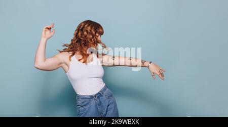 Donna dai capelli rossi che ballava mentre si era in piedi in uno studio. Donna che si diverte in abbigliamento casual. Foto Stock
