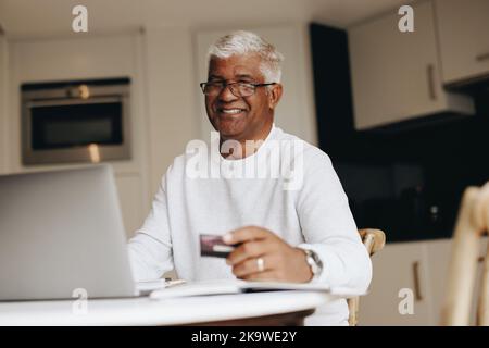 Felice uomo anziano sorridente alla macchina fotografica mentre acquista in linea a casa. Uomo maturo che usa la sua carta di credito per effettuare un ordine online su un computer portatile. Allegro Foto Stock