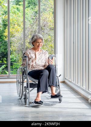 Donna anziana asiatica felice si siede in sedia a rotelle, salutando, sorridendo e guardando lo schermo del telefono cellulare intelligente, stile verticale. Le donne anziane incontrano fa Foto Stock