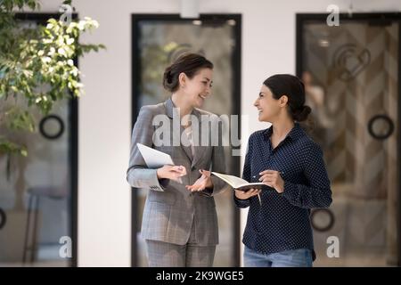 Due attraenti donne d'affari multietniche che parlano in piedi nel corridoio dell'ufficio Foto Stock