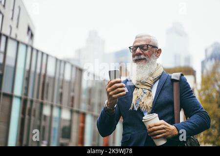 Uomo anziano di hipster di affari che usa il telefono mobile mentre cammina per lavorare con la città in background - faccia di fuoco Foto Stock