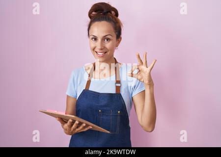 Bruna donna che indossa cameriera professionale grembiule tenere appunti facendo segno ok con le dita, sorridendo amichevole gesturing eccellente simbolo Foto Stock