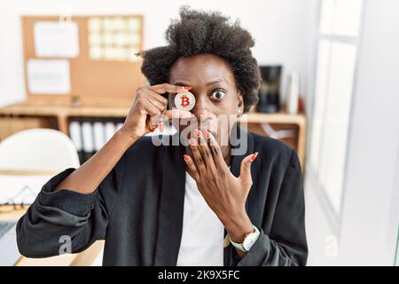 Giovane donna africana che tiene il bitcoin sugli occhi in ufficio che copre la bocca con la mano, scioccato e impaurito per errore Foto Stock