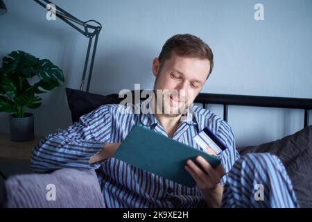 Giovane uomo attraente che usa un tablet seduto a letto la mattina facendo acquisti online. Maschio caucasico che utilizza il touchpad per lo shopping con carta di credito in mano. Immagini di alta qualità Foto Stock
