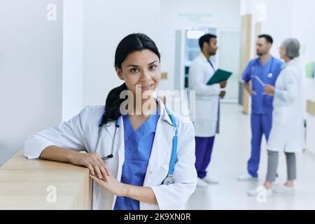 Medico asiatico positivo donna in piedi con stetoscopio nel corridoio moderno ospedale sullo sfondo di assistenti medici di lavoro, medici e NUR Foto Stock