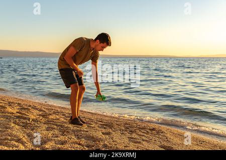 Giorno della terra. Pulire la spazzatura sulla costa adriatica. Un ragazzo che raccoglie una bottiglia di plastica sulla spiaggia. Tramonto. Il concetto di conservazione dell'ecologia Foto Stock