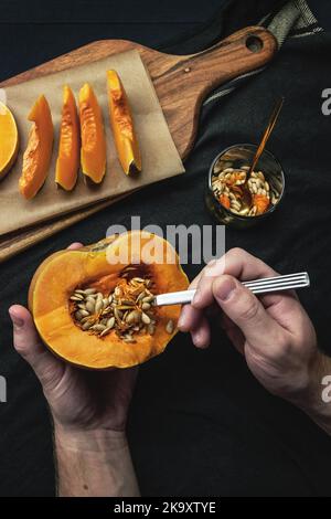 Mani maschili che rimuovono i semi con il cucchiaio da una zucca tagliata di fresco su fondo nero Foto Stock