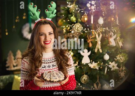 Natale. Sorridente elegante casalinga di 40 anni con biscotti di Natale in maglione di Natale tradizionale vicino all'albero di Natale nella casa moderna. Foto Stock