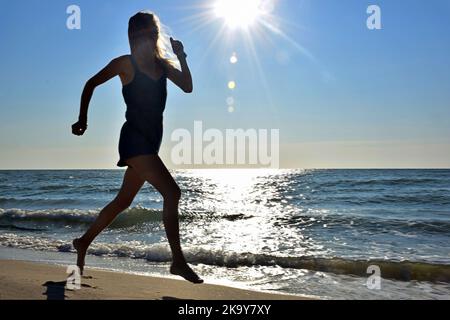 Giovane donna bionda di razza bianca corre felice lungo la spiaggia della riva in tute denim Foto Stock