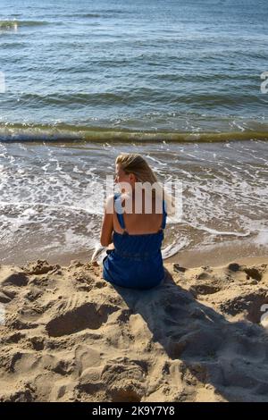 Una giovane donna siede sulla sabbia sulla spiaggia vicino al mare e guarda in lontananza. Vista posteriore Foto Stock