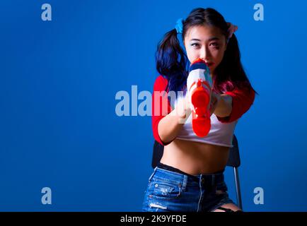 Giovane donna in costume di Harlequin seduto e puntando pistola d'acqua alla telecamera con Copy Space a sinistra in isolato sfondo blu Foto Stock