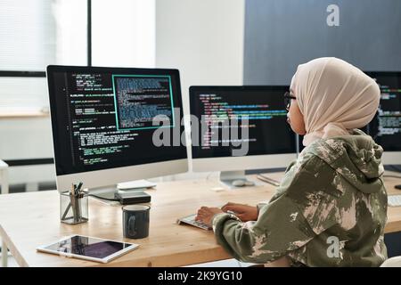 Vista laterale di una giovane donna d'affari musulmana in rete davanti allo schermo del computer con informazioni codificate dal posto di lavoro Foto Stock