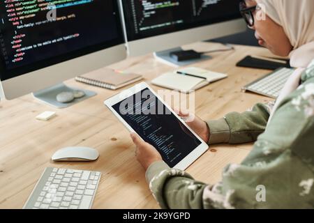 Primo piano di una giovane programmatore musulmana che guarda i dati codificati sullo schermo del tablet mentre si siede davanti ai monitor del computer Foto Stock