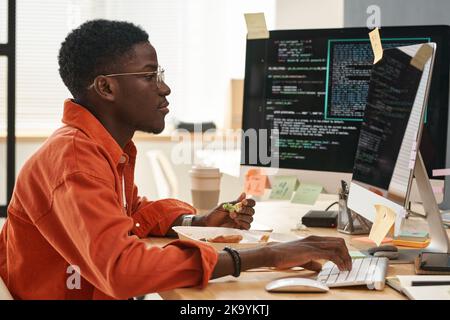 Vista laterale del giovane uomo nero in casualwear intelligente mangiare cibo sano per la prima colazione mentre si siede sul posto di lavoro di fronte allo schermo del computer Foto Stock