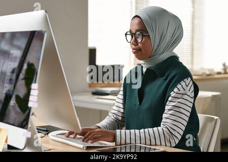 Giovane donna musulmana fiduciosa intern o designer in occhiali, hijab e casualwear di rete di fronte al monitor del computer Foto Stock