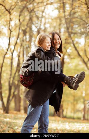 Gioire giovane donna cavalcare sul retro piccola ragazza vista laterale. Felice madre e figlia in abiti neri casual divertirsi e giocare nel parco d'autunno dorato Foto Stock