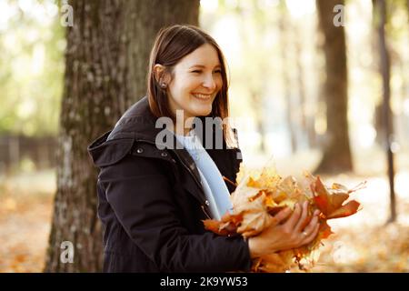 Ritratto orizzontale di donna sorridente in giacca nera che getta su manciata di foglie gialle asciutte su fondo di foresta dorata. Felice ragazza a piedi e. Foto Stock