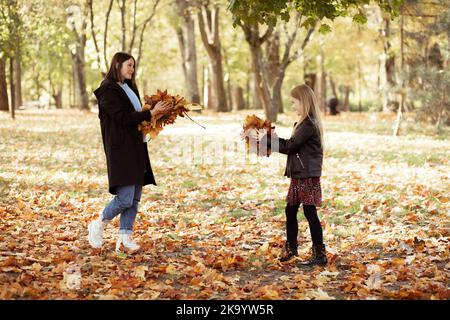 Giovane donna allegra e bambina buttare su mazzo di foglie gialle asciutte. Felice madre e figlia in abiti neri casual divertirsi e giocare Foto Stock