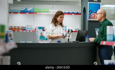 Il farmacista parla con il cliente senior delle pillole cardiologiche, della scansione della scatola dei farmaci e del lavoro presso la farmacia. Lavoratore medico che spiega la prescrizione di vitamine e integratori a farmacia. Foto Stock