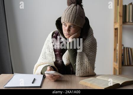 arrabbiata donna congelata vestita calorosamente in una casa fredda seduta a un tavolo con telefono cellulare, problemi di riscaldamento, crisi energetica. niente calore Foto Stock