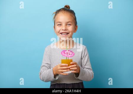 Allegra bambina beve succo d'arancia e ride, si trova su sfondo isolato blu. Foto Stock