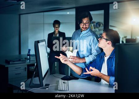 Discutere il lavoro in corso. Un gruppo di programmatori che lavorano insieme su un computer in ufficio durante la notte. Foto Stock