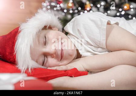 Natale sogni di un bambino. Un bambino in un cappello di Babbo Natale dorme a letto vicino a un albero di Natale con le luci di Natale, in attesa del regalo di Babbo Natale. Foto Stock