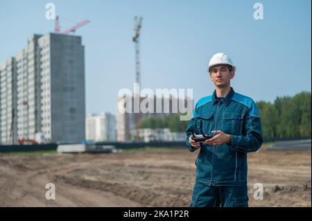 Un uomo in un casco e tute controlla un drone in un cantiere. Il costruttore esegue la supervisione tecnica. Foto Stock