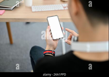 Vista posteriore, giovane uomo asiatico seduto alla sua scrivania e utilizzando il suo smartphone per parlare con gli amici. Telefono schermo bianco mockup Foto Stock