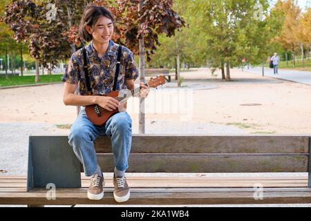 Ragazzo asiatico sorridente e suonante la chitarra acustica Ukulele seduta su una panchina. Foto Stock