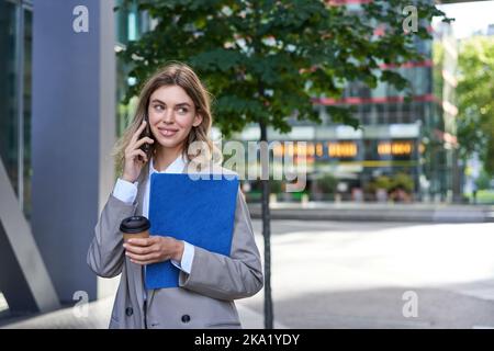 Occupato donna d'affari beve caffè, parla sul telefono cellulare, tiene documenti per il lavoro, passeggiate nel centro della città vicino ufficio Foto Stock