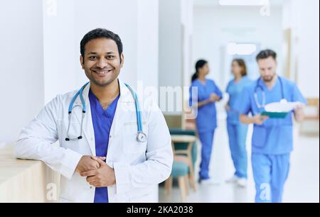 Medico indiano sorridente in piedi nel corridoio di un ospedale moderno sullo sfondo di assistenti medici e infermieri funzionanti. istituzione medica Foto Stock