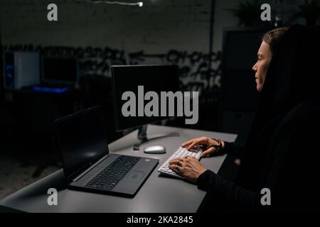 Vista laterale di un pericoloso hacker che indossa una tastiera con cappuccio che interrompe la password, guardando lo schermo del computer portatile e del desktop. Foto Stock