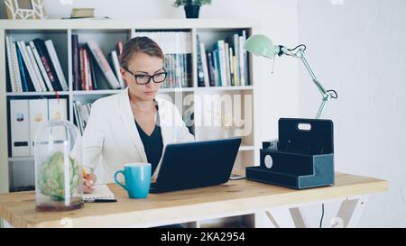 Attraente giovane donna sta lavorando in un moderno ufficio seduto al tavolo, utilizzando il computer portatile e la scrittura in notebook. Sono visibili scaffali con carte, piante verdi e libri Foto Stock