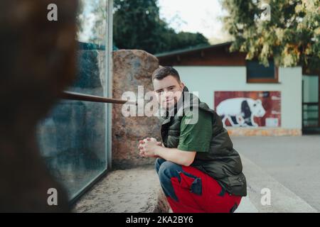 Giovane uomo con sindrome di Down, che riposa dopo aver pulito il recinto degli animali nello zoo. Concetto di integrazione delle persone con disabilità nella società. Foto Stock