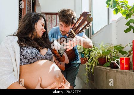 giovane coppia latina incinta romantica, l'argentino caucasico con la sua moglie brasiliana stanno godendo fare la musica e cantare rilassati a casa, avendo Foto Stock
