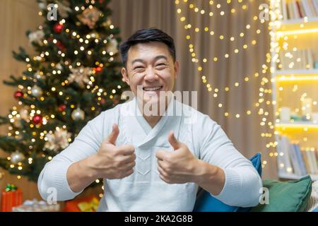 L'uomo asiatico sul Natale guarda nella macchina fotografica di fotoricettore e sorride mostra le sue barrette in su affermativamente, l'uomo si siede nel paese sul divano vicino all'albero di Natale per il nuovo anno. Foto Stock