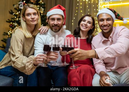 Ritratto di un gruppo di ospiti diversi a casa per il nuovo anno, amici che festeggiano il Natale guardando la macchina fotografica e sorridendo tenendo bicchieri di vino, seduti sul divano nel salone del cinema. Foto Stock