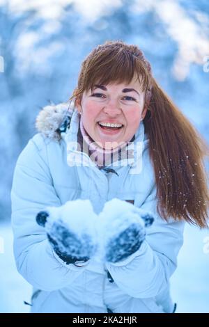 Ritratto di una giovane donna che soffia neve dalle palme in una foresta invernale innevata. La ragazza è entusiasta della neve. Foto Stock