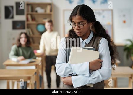 Offese l'allievo della scuola secondaria con libri in piedi davanti alla macchina fotografica con l'espressione triste contro due scolari che la bullonano Foto Stock